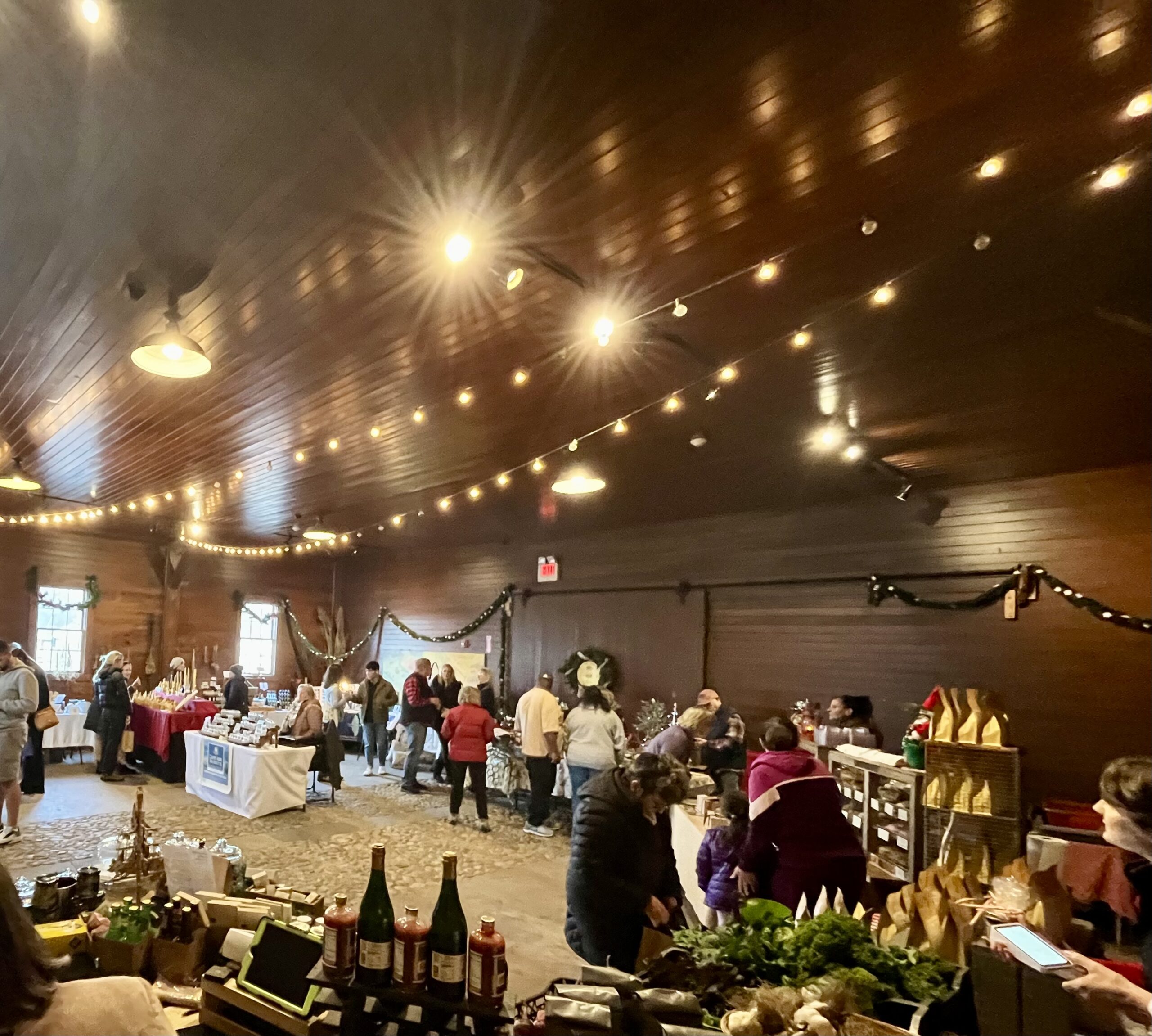 the inside of a barn, set up for a holiday market with lights and evergreens