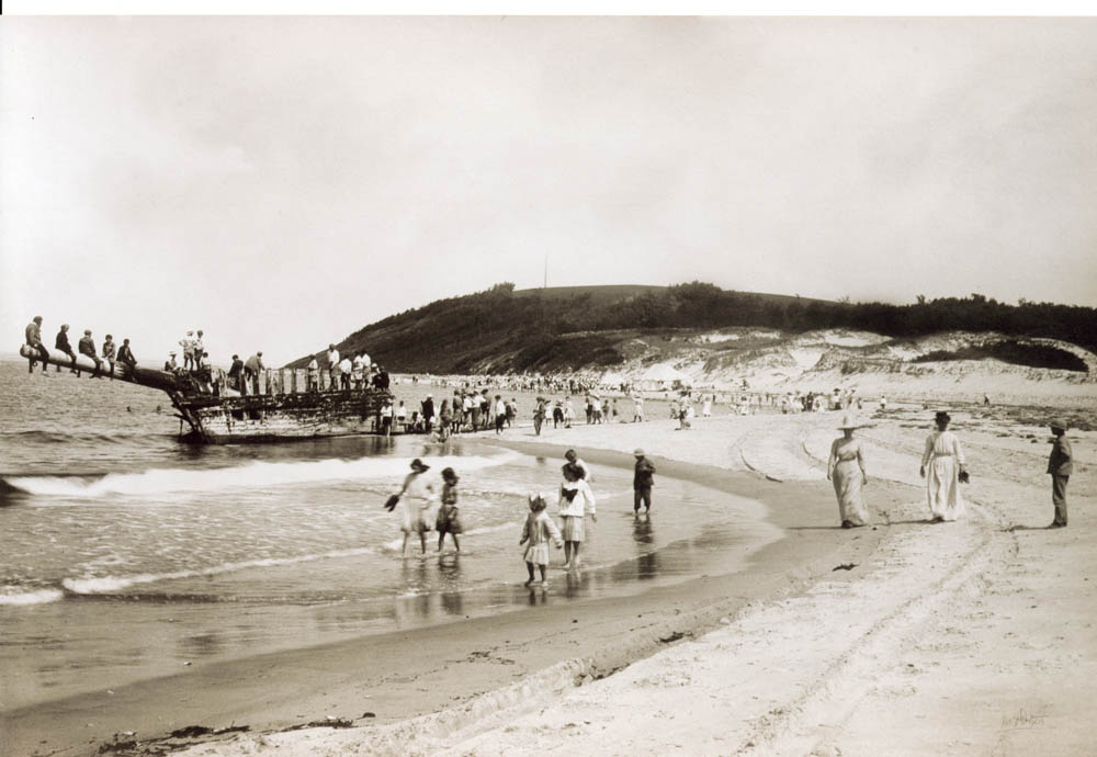 A black and white photograph of a beach with many people in the foreground and people sitting on the hull of a shipwreck in the background.