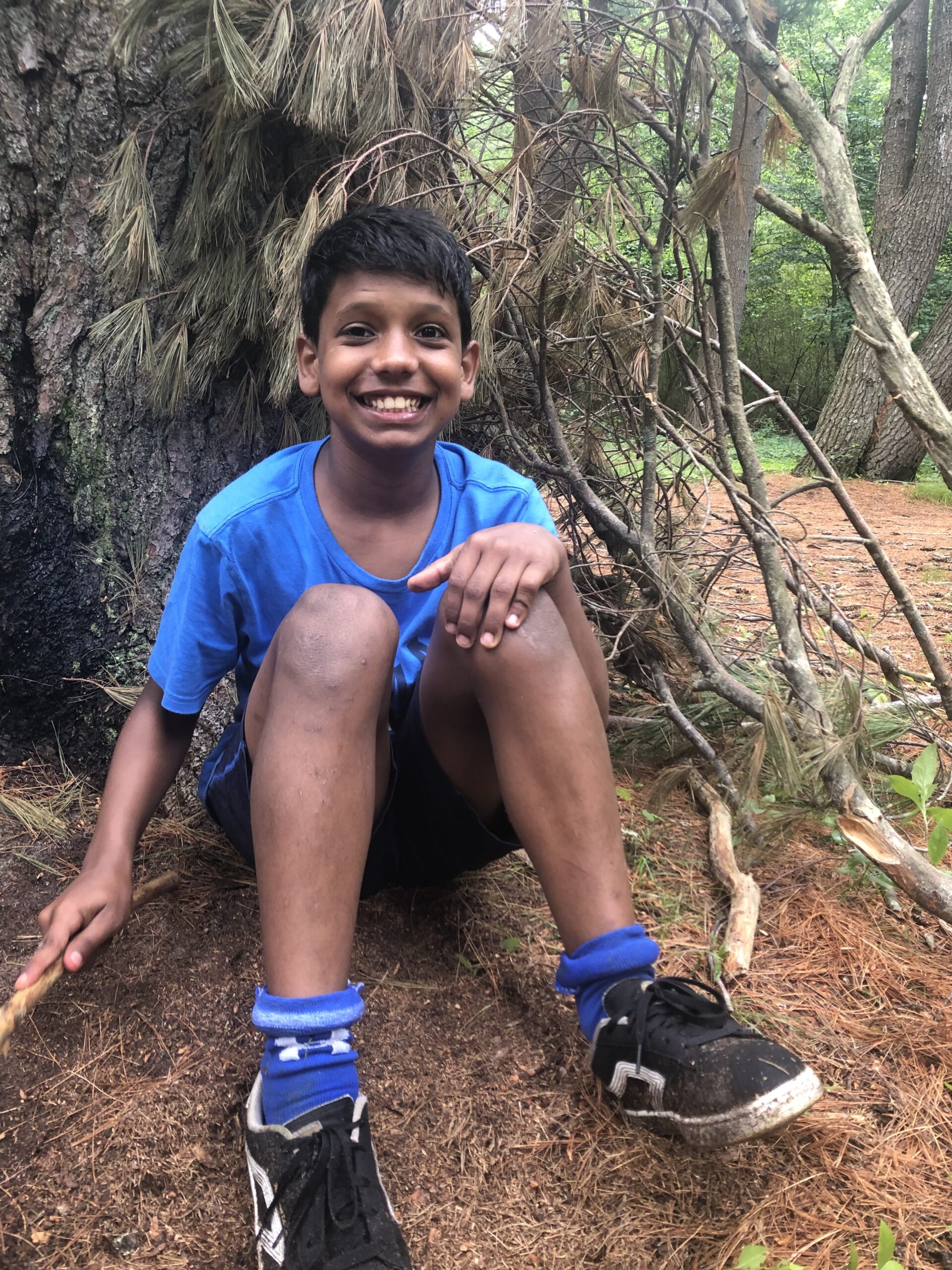 A smiling boy sitting on the ground beneath a tree wearing a blue shirt and hiking shoes