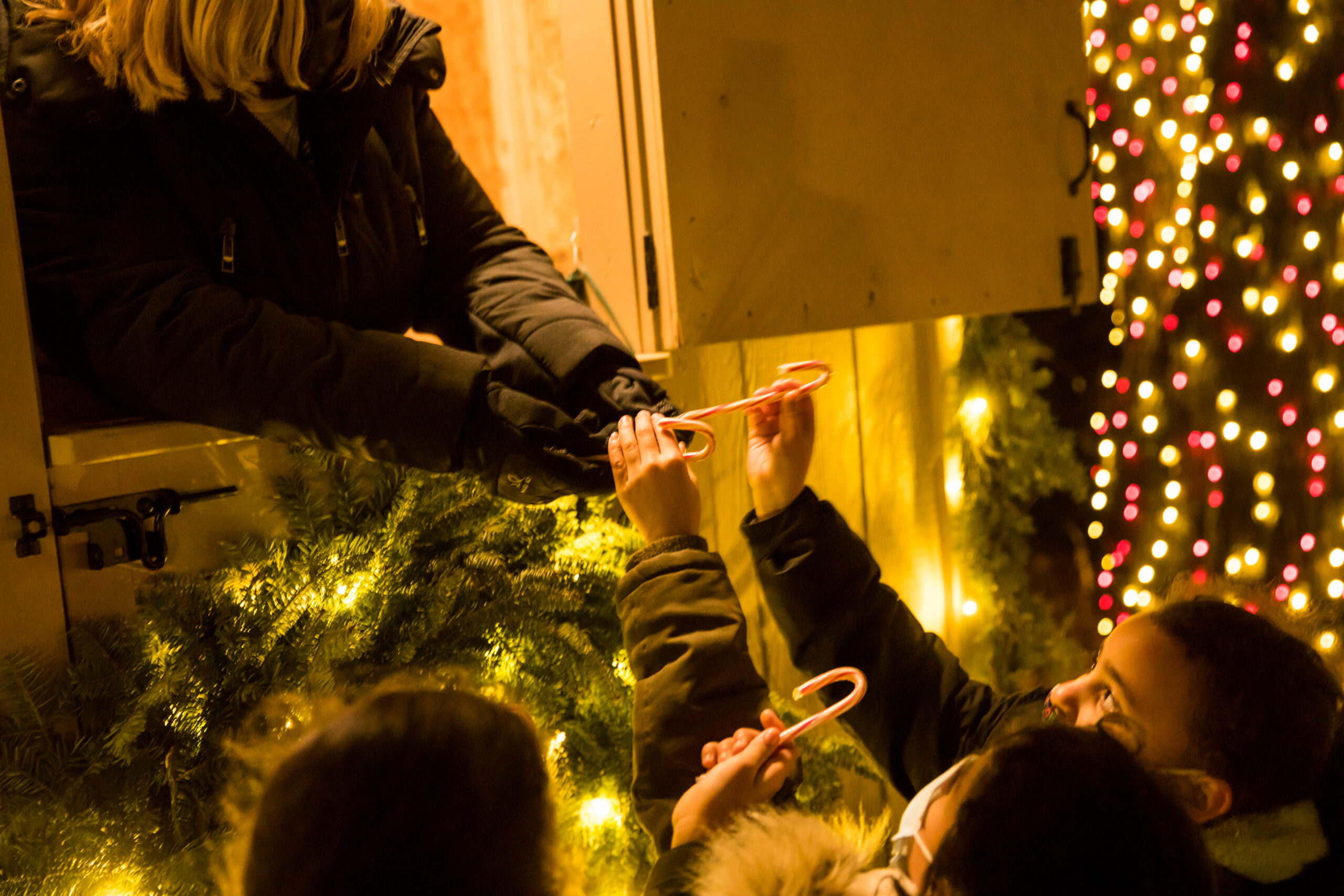 Four children standing outdoors beneath a window receiving candy canes from someone inside, surrounded by holiday lights at Winterlights