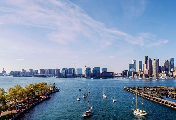 Blue water with boats and green land on either side with the Boston skyline and blue sky in the background