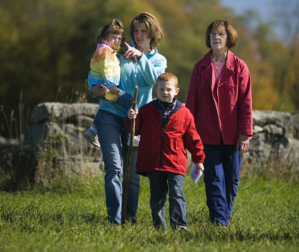 Two women hiking in a green field with two children during the day with fall foliage in the background