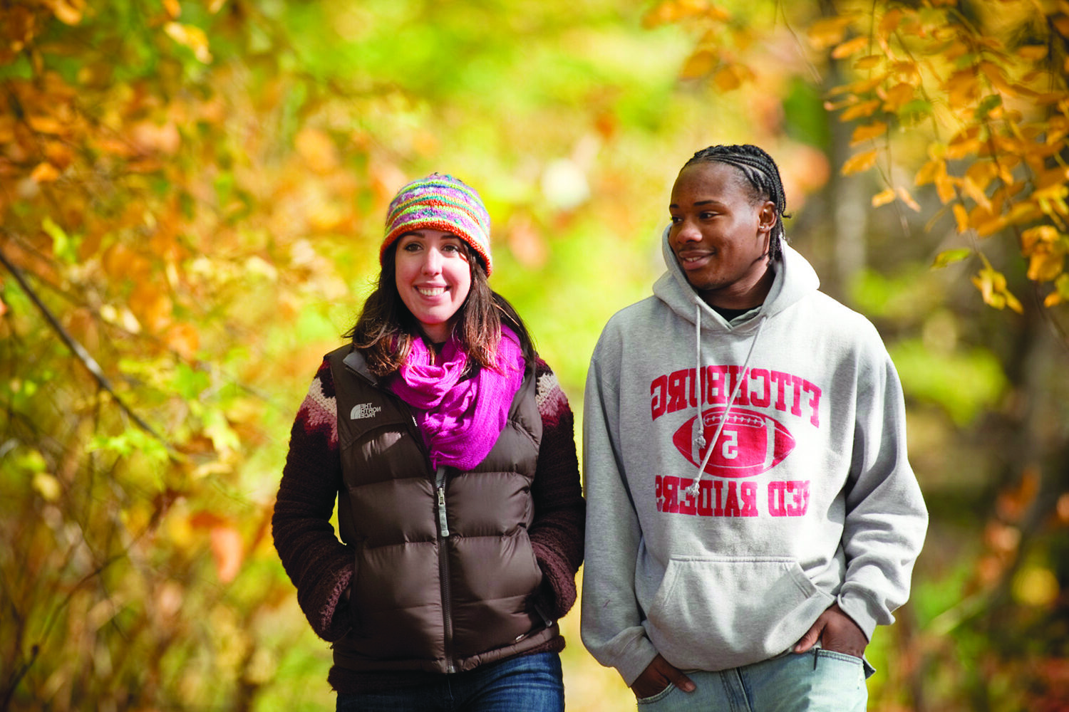 Two smiling people walking down a path outdoors surrounded by fall foliage