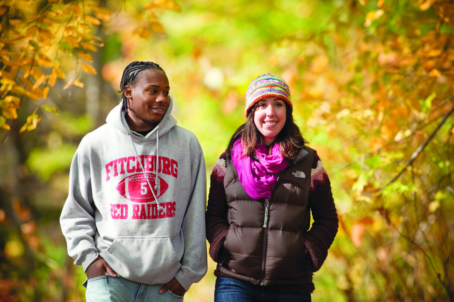Two smiling people walking down a path outdoors surrounded by fall foliage