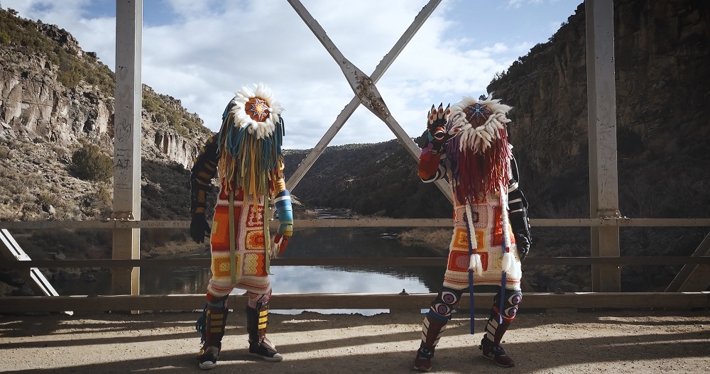 two figures in Indigenous dress stand on a bridge with mountains in the background