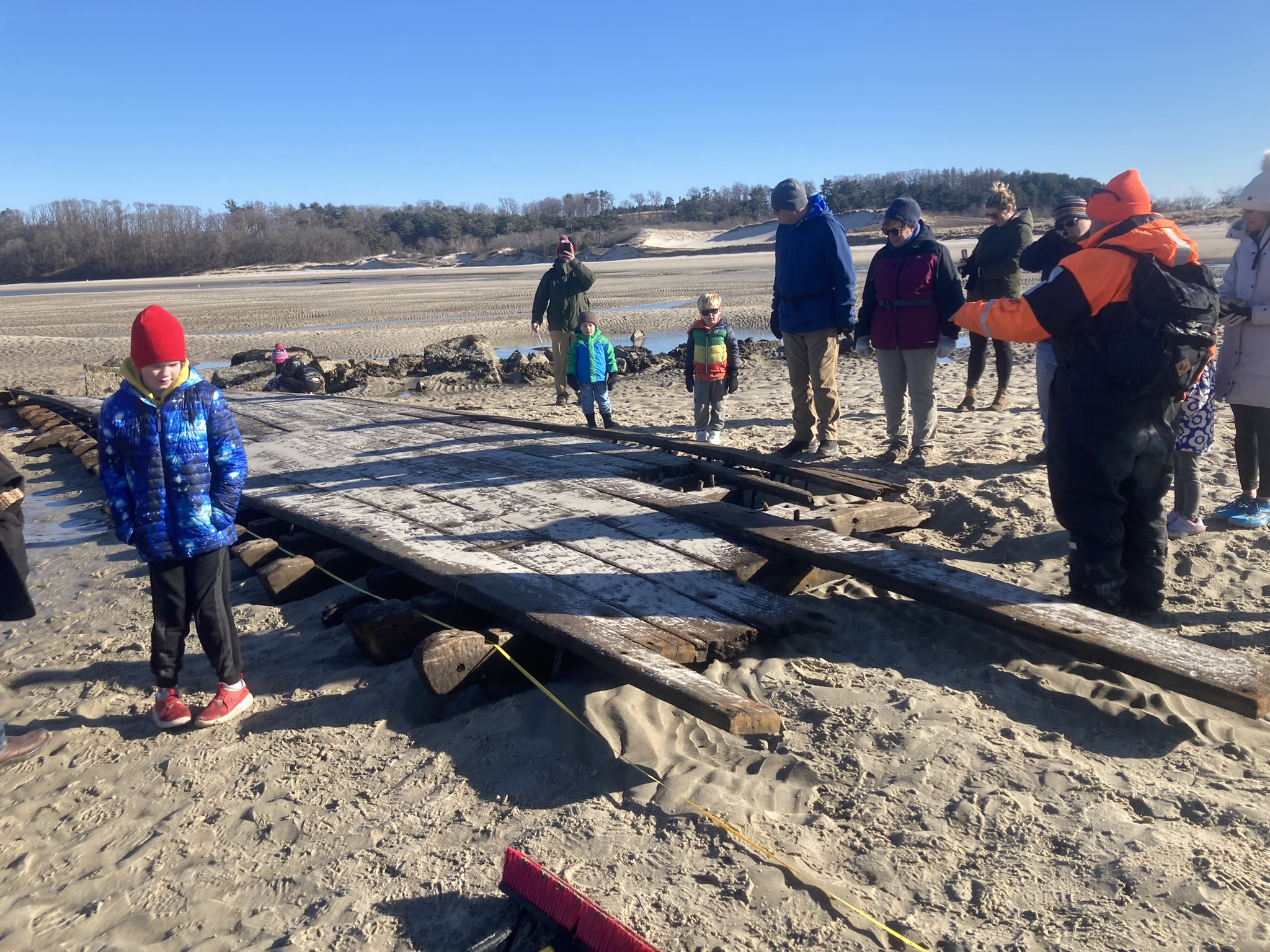 Several people look at an uncovered part of a shipwreck that rests on a sandy beach.