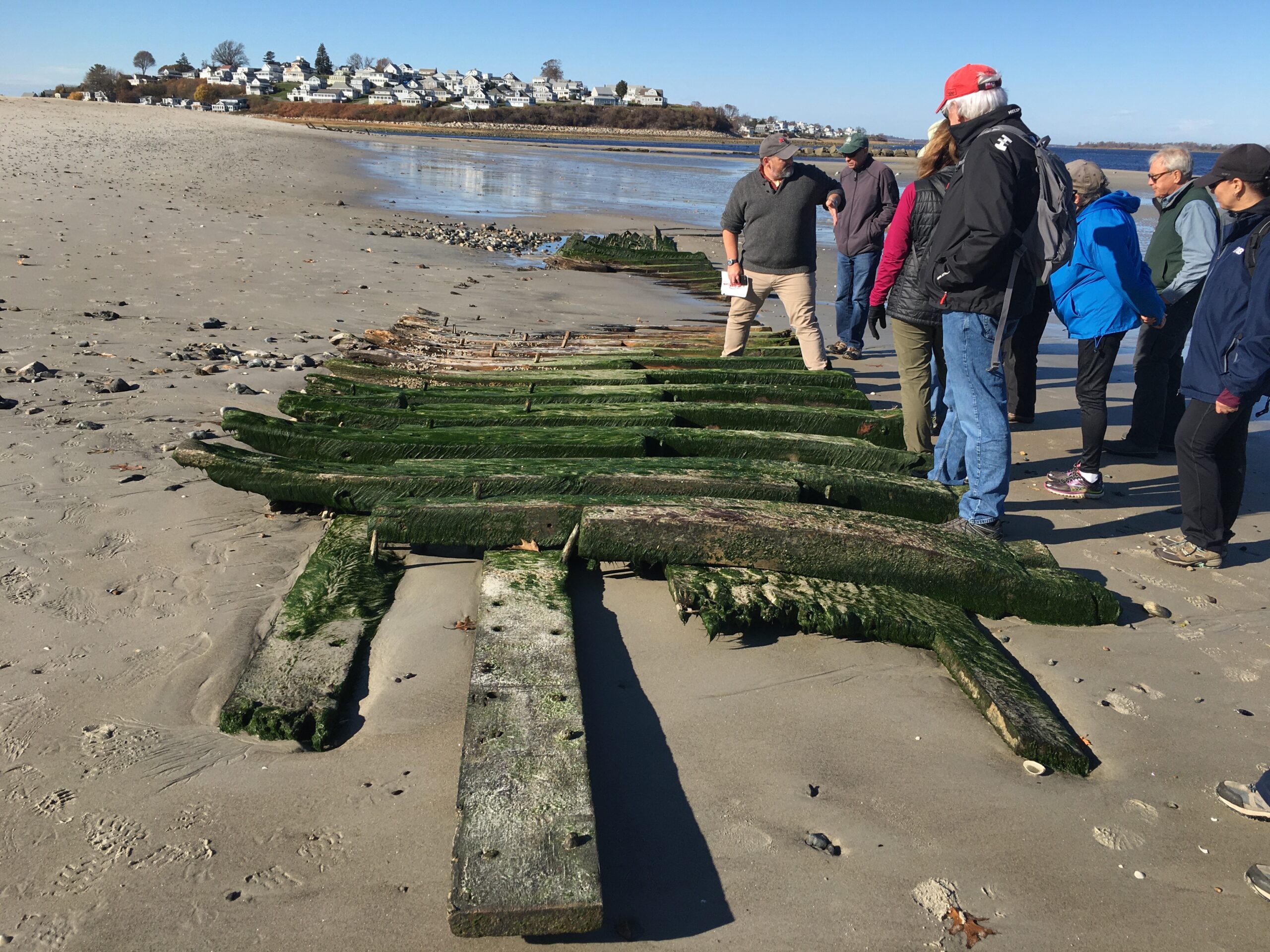 Several people look at a piece of Ada K. Damon, a shipwreck located on a sandy beach.