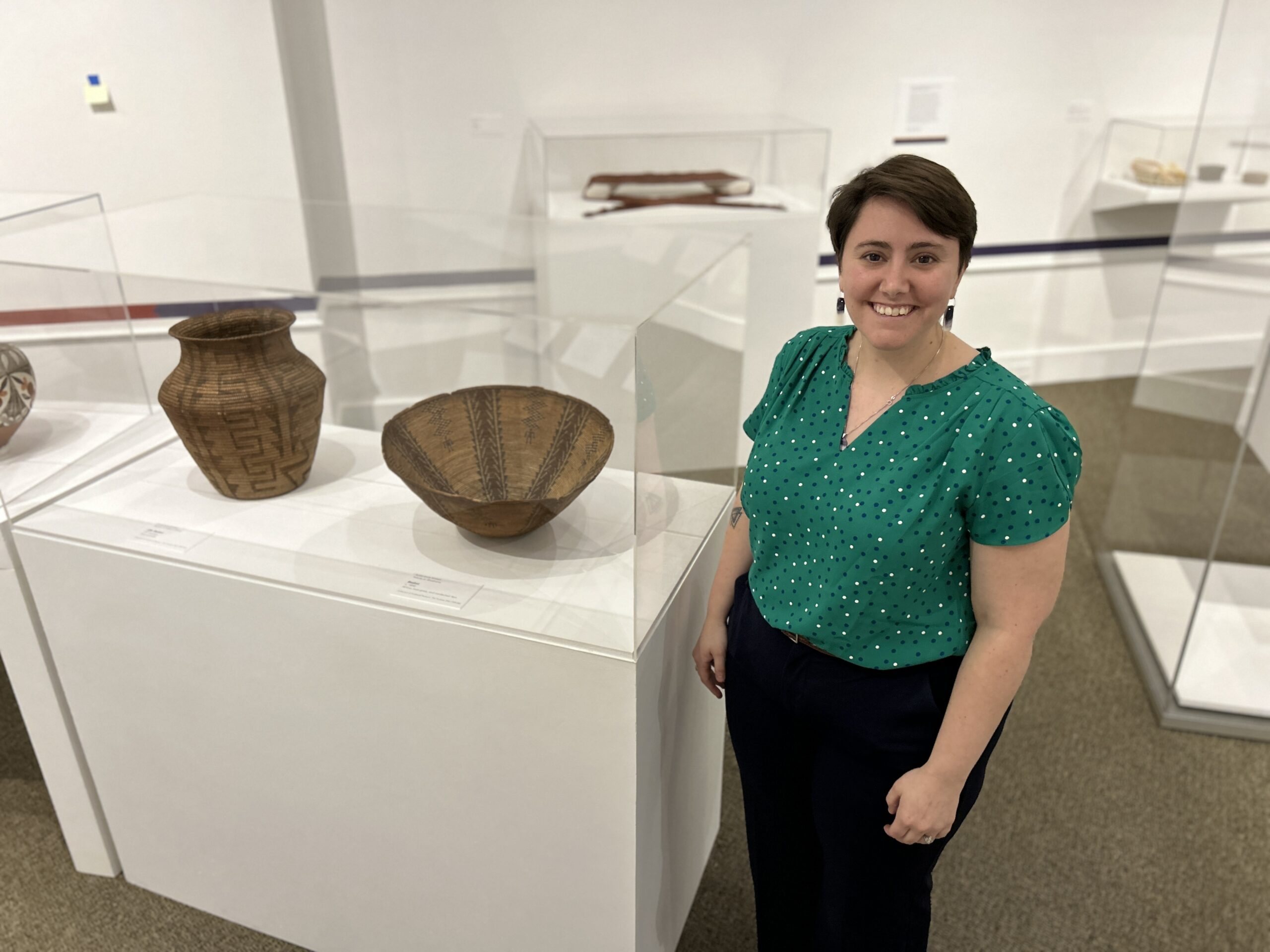 a smiling woman with short hair and a green shirt stands next to native baskets on display