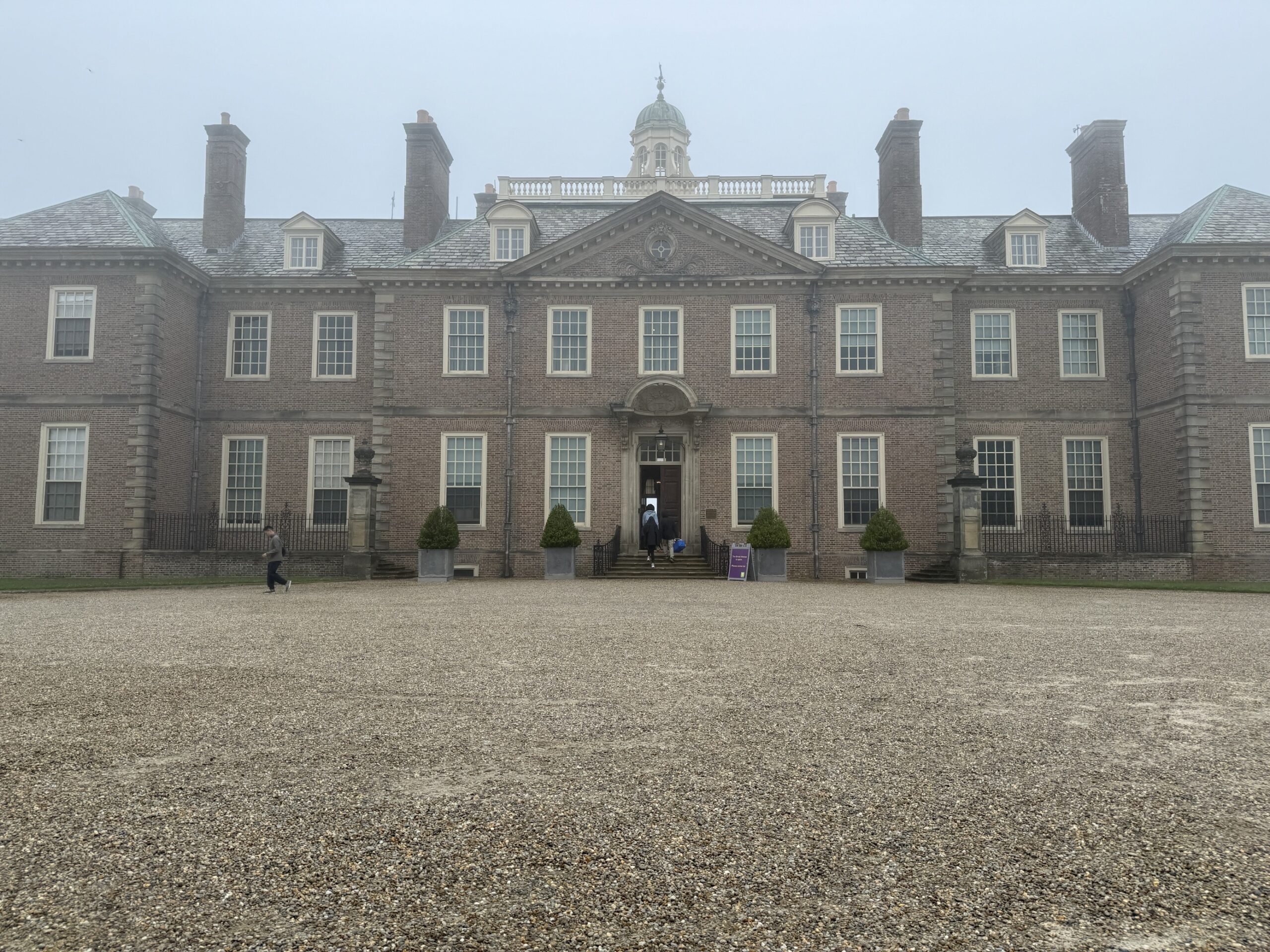 A large estate made of stone is pictured with a pebbled drive on a foggy and grey day.
