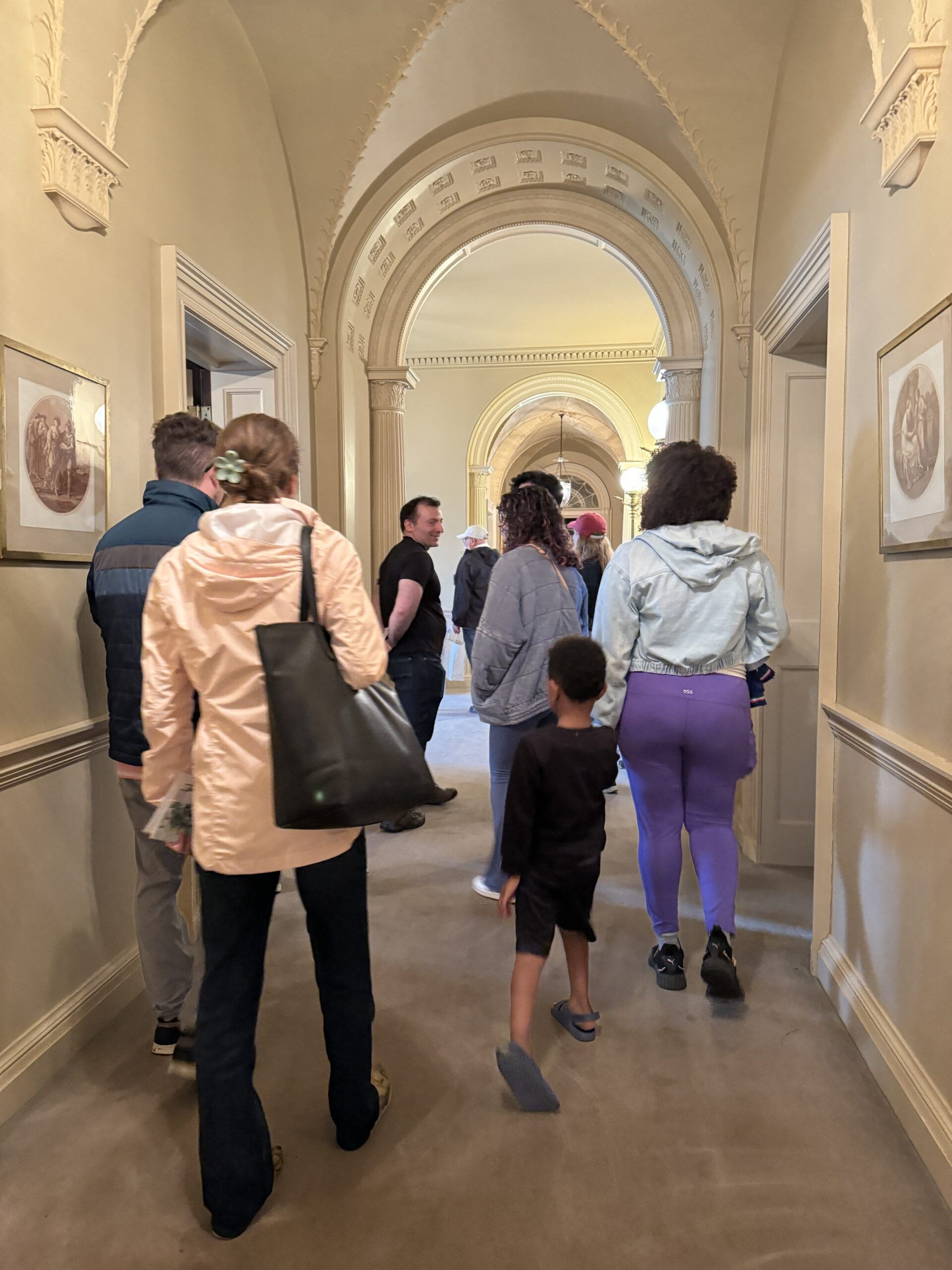 Several people with their backs turned walk the halls of the Crane Estate. They are walking through an arched cream-colored hallway with art on both sides.