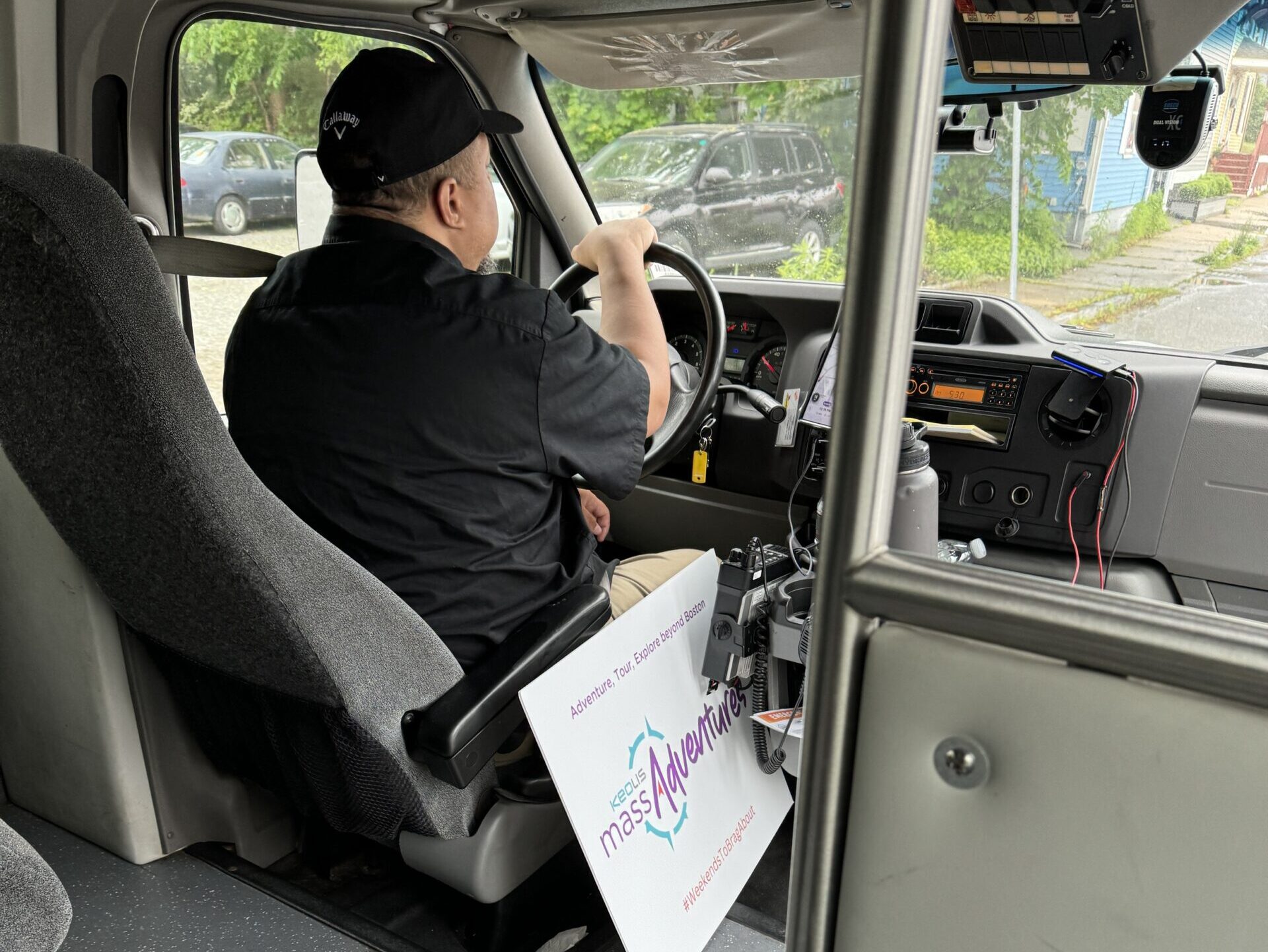 A man sits with his back to the camera in a black t-shirt and black baseball hat. He has one hand on a steering wheel, driving a bus.
