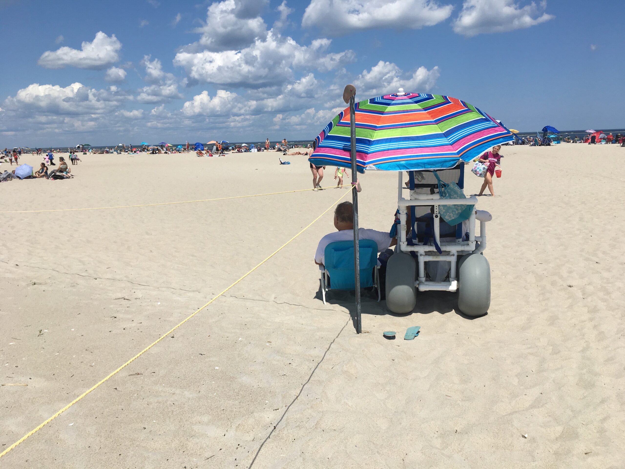Two people sit with backs to the camera on the beach under an umbrella. One person is in a beach wheelchair and the other is sitting in a beach chair.