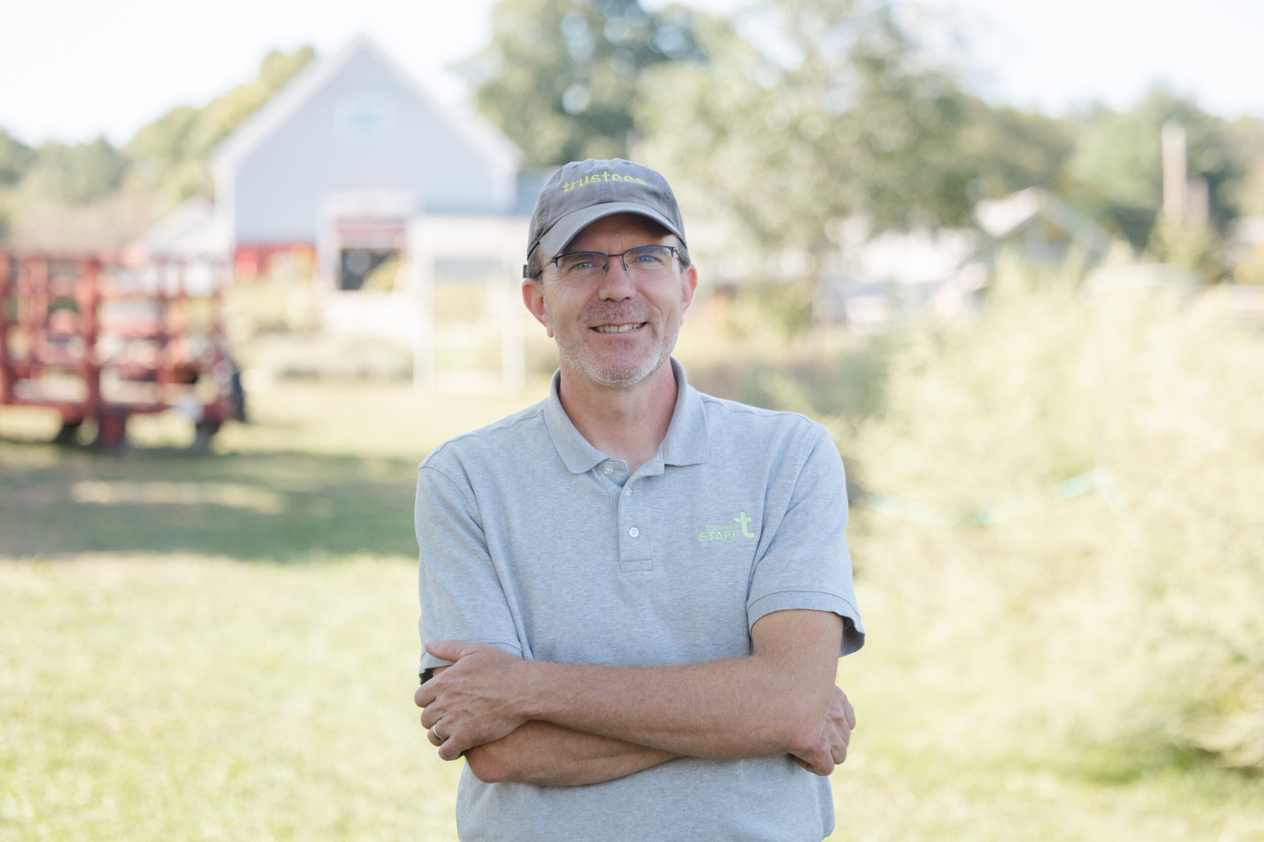 A man smiles at the camera wearing a baseball hat and a polo shirt with the Trustees logo, crossing his arms