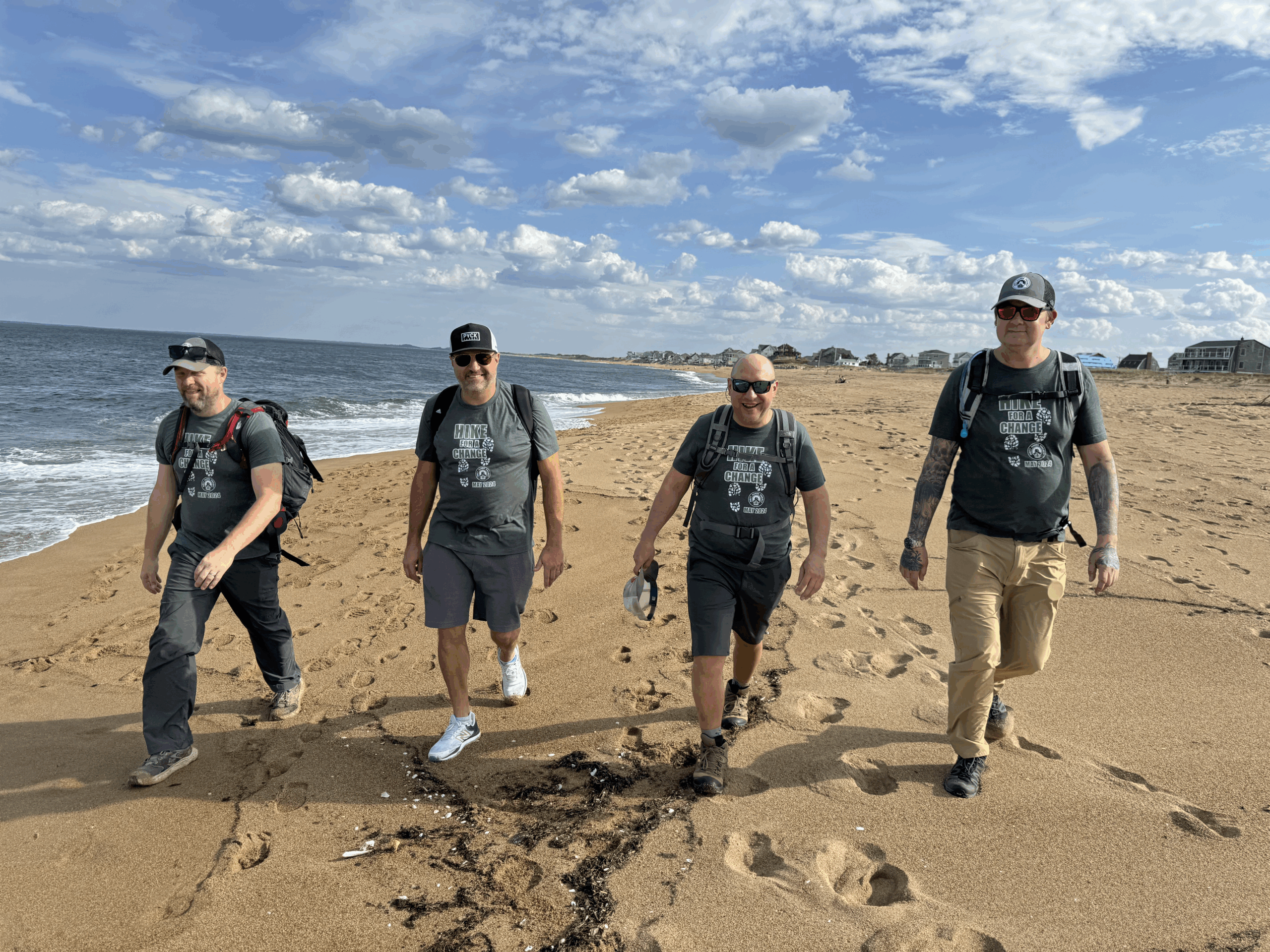 Four people smile as they walk towards the camera on a beach on a sunny day.
