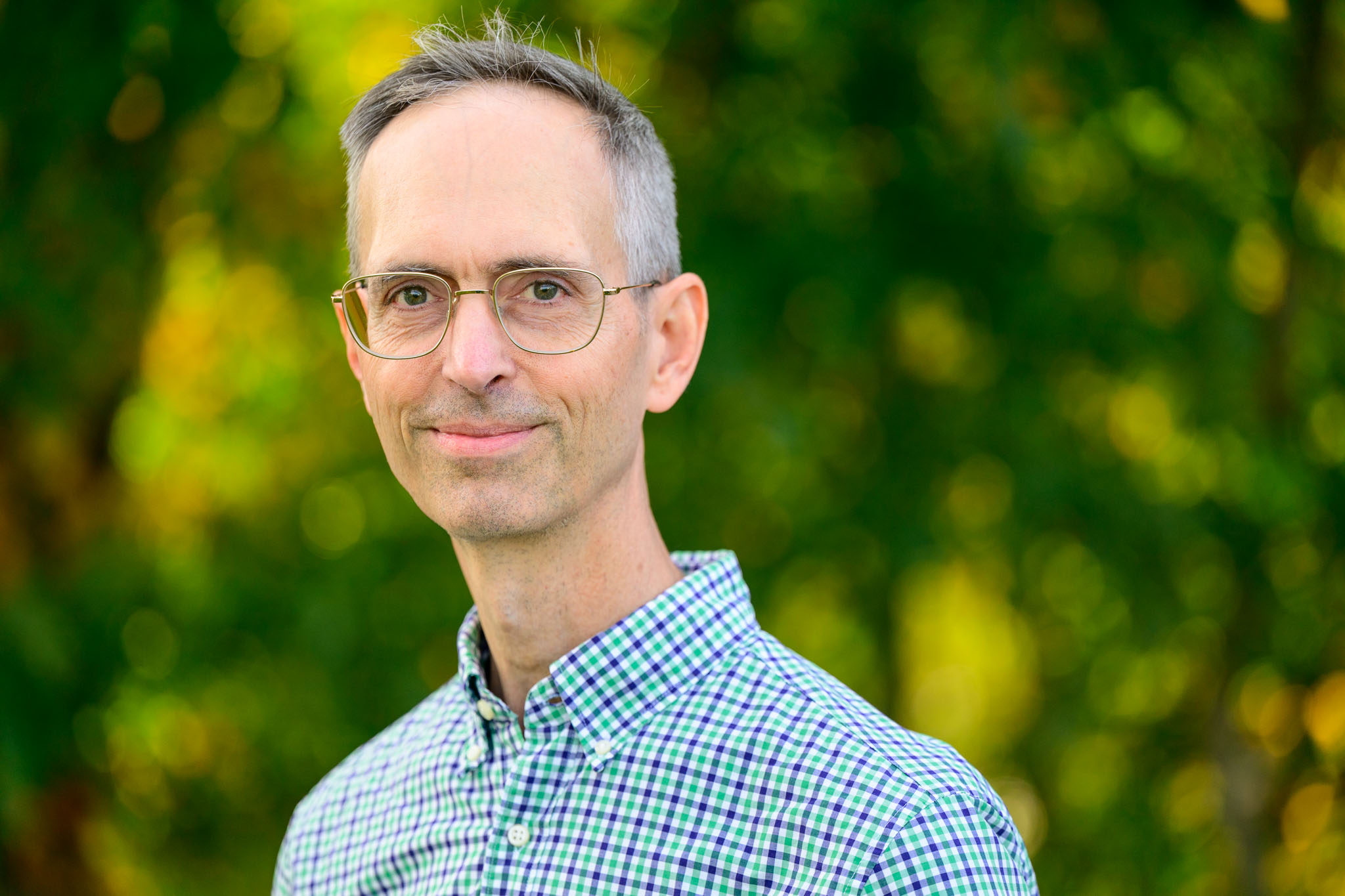 A white man with short grey hair and glasses softly smiles at the camera