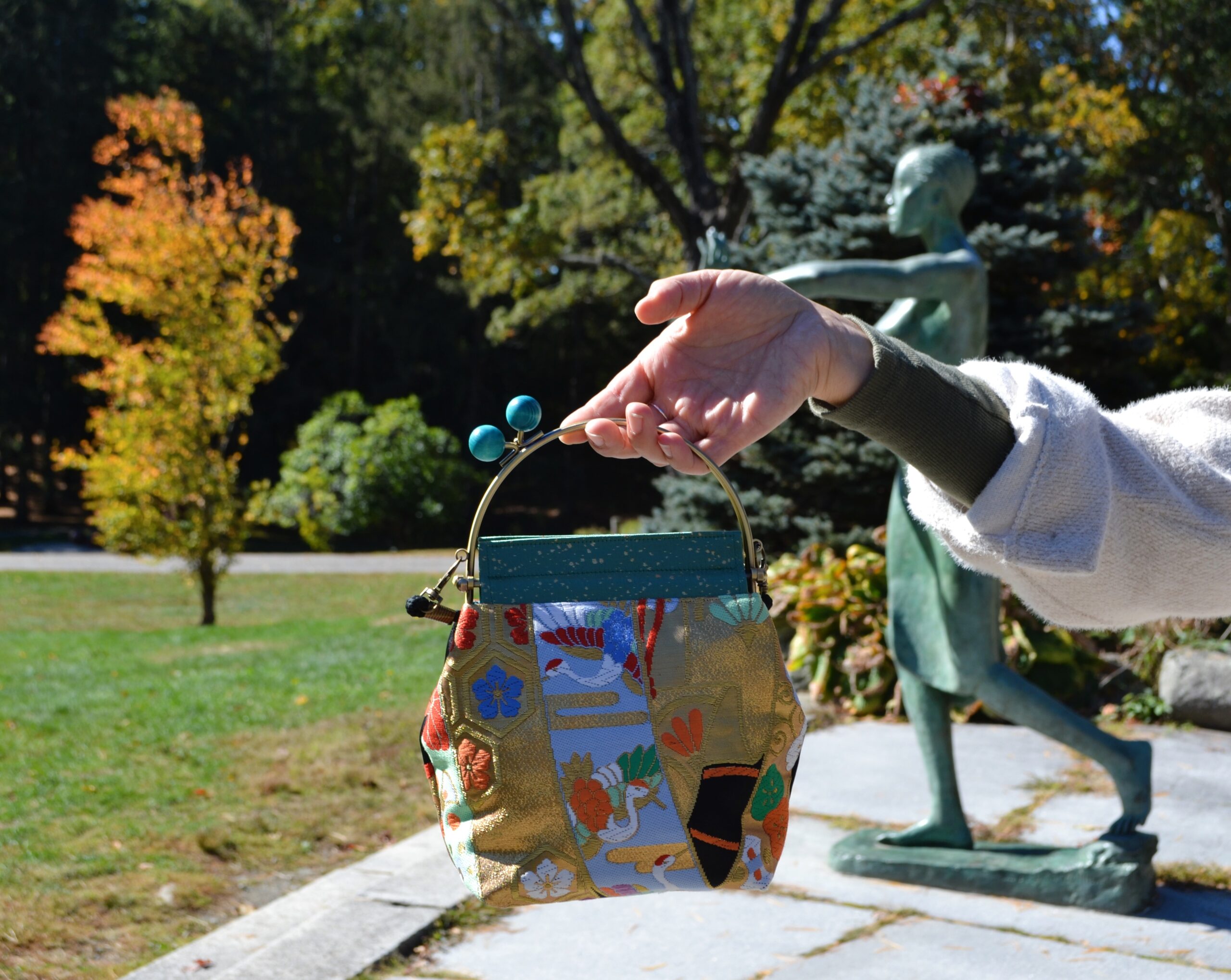 A multi-colored small bag made out of kimono and obi is held up by a hand in front of a green sculpture