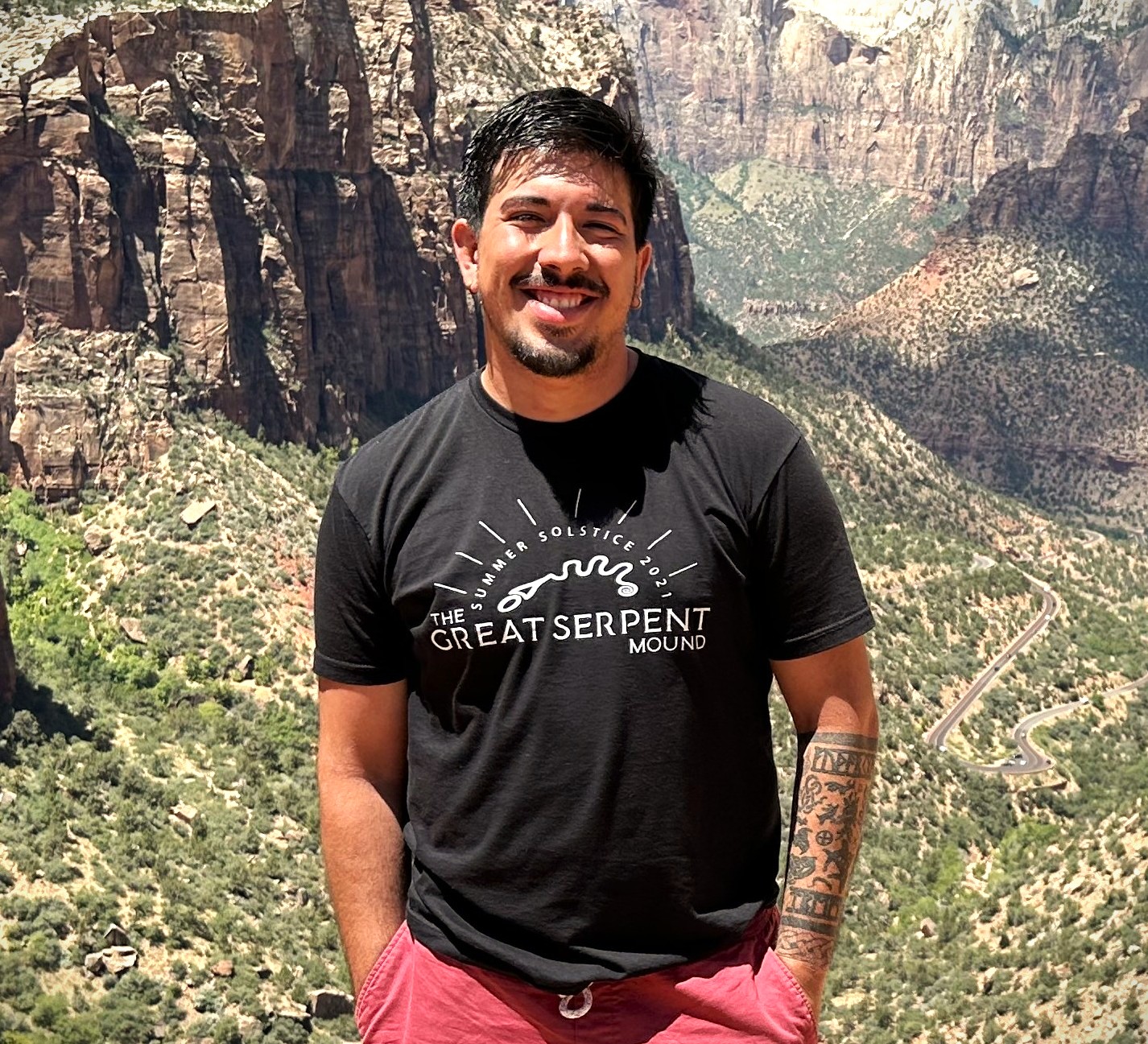 A man in a dark t shirt and red shorts smiles at the camera at Zion National Park.
