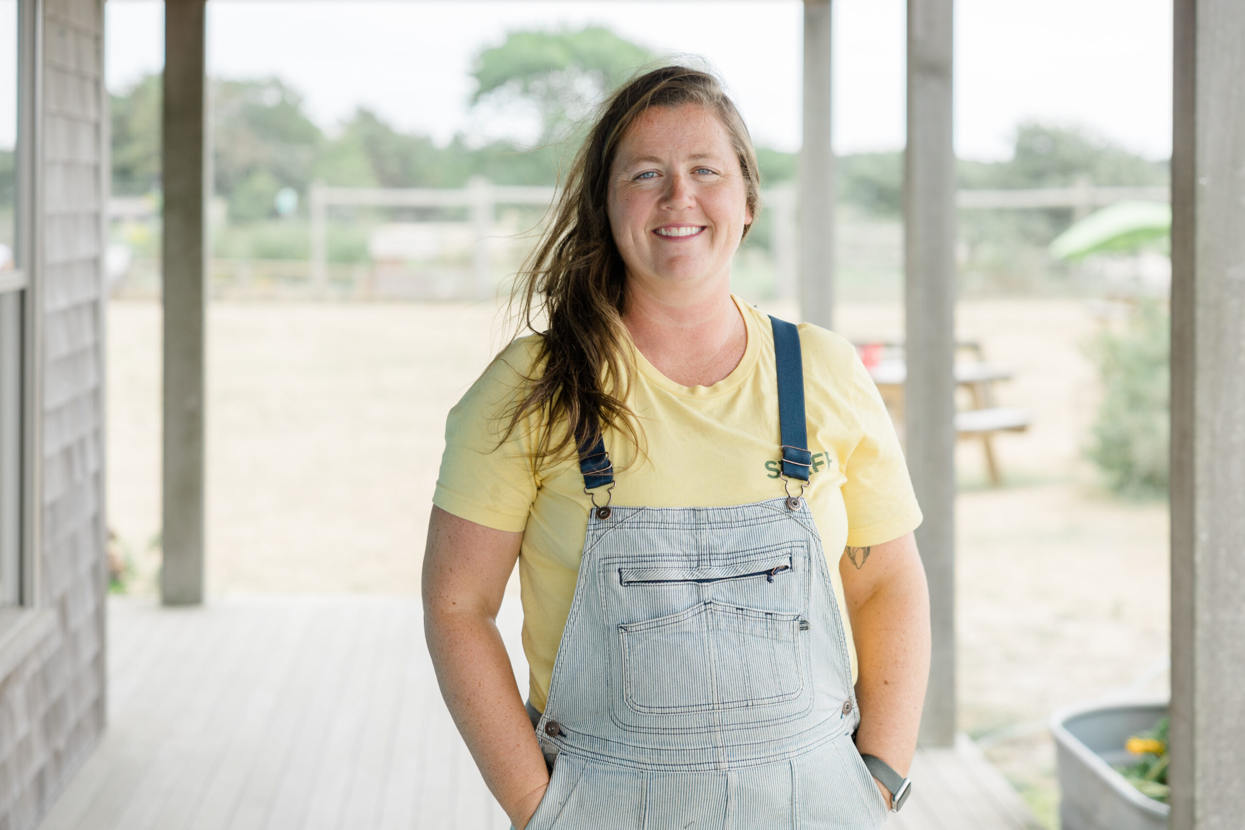 A woman smiles at the Camera with long brown hair, a yellow shirt, and overalls.