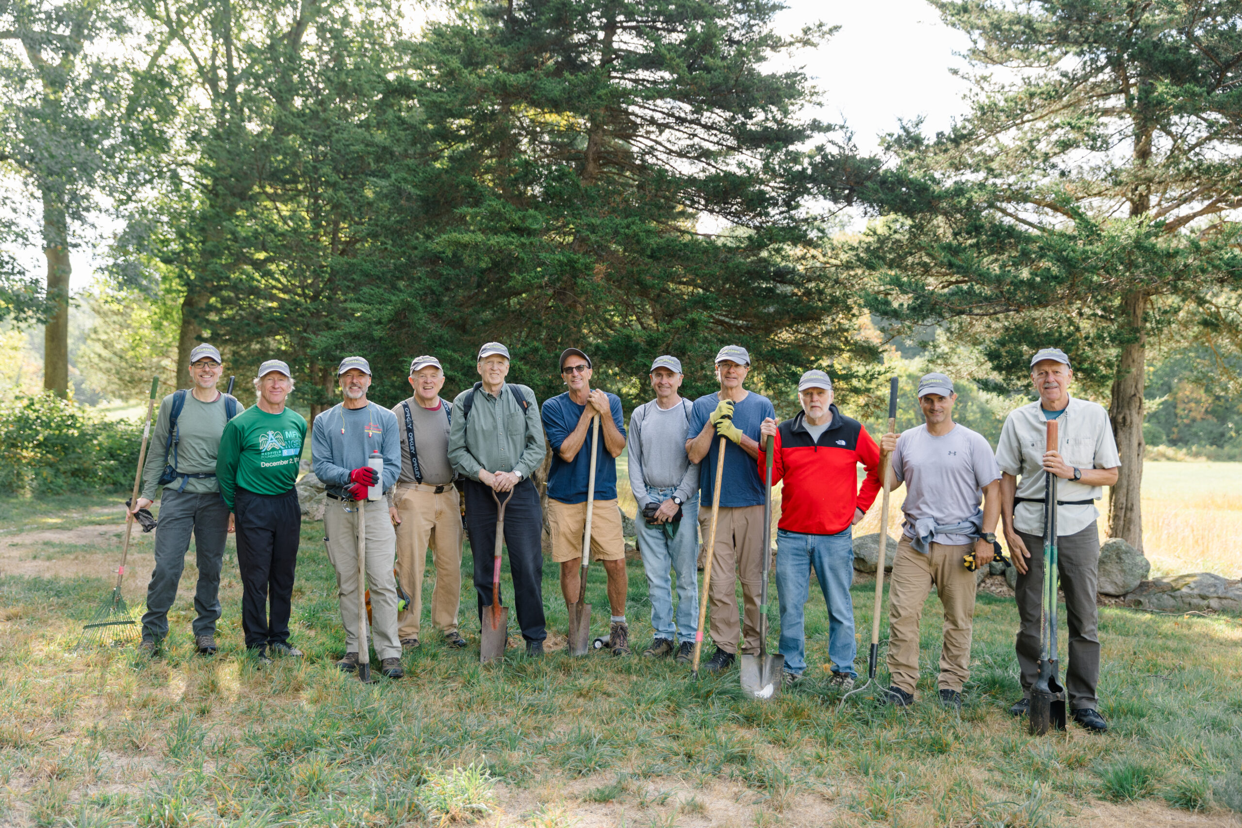 A group holding various landscaping tools smiles at the camera in a forest clearing.