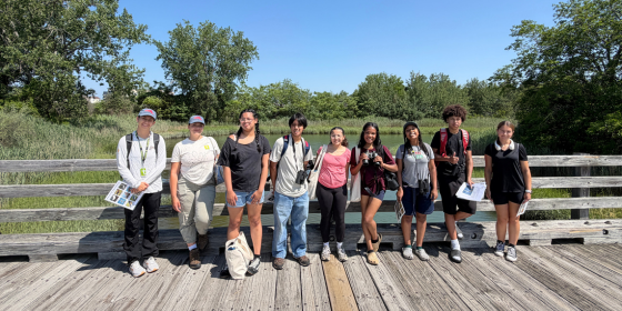 A group of teens smiles at the camera on a boardwalk