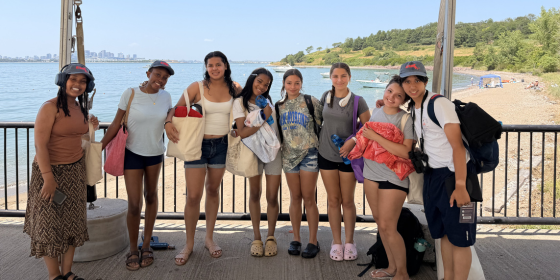 A group of teens poses to smile for the camera in front of a beach